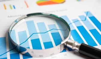 A magnifying glass placed over a printed chart showing blue bar graphs and a blue line graph, symbolizing market research