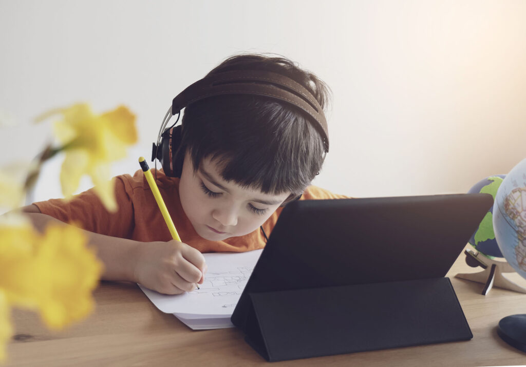 child writing in a notebook with an iPad in front of them